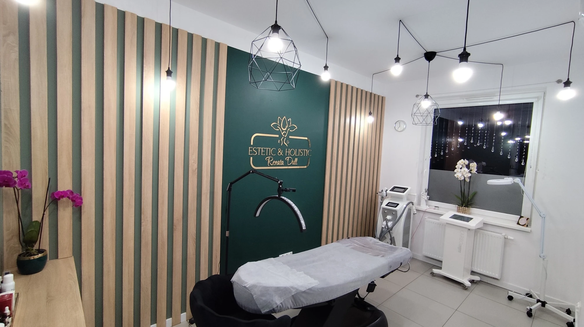 Gabinet zabiegowy RD ESTETIC and HOLISTIC w panoramicznym ujęciu - zielona szałwiowa ściana z gold logo, drewniane lamele, geometryczne czarne lampy, białe łóżko zabiegowe, orchidee, sprzęt kosmetologiczny przy oknie